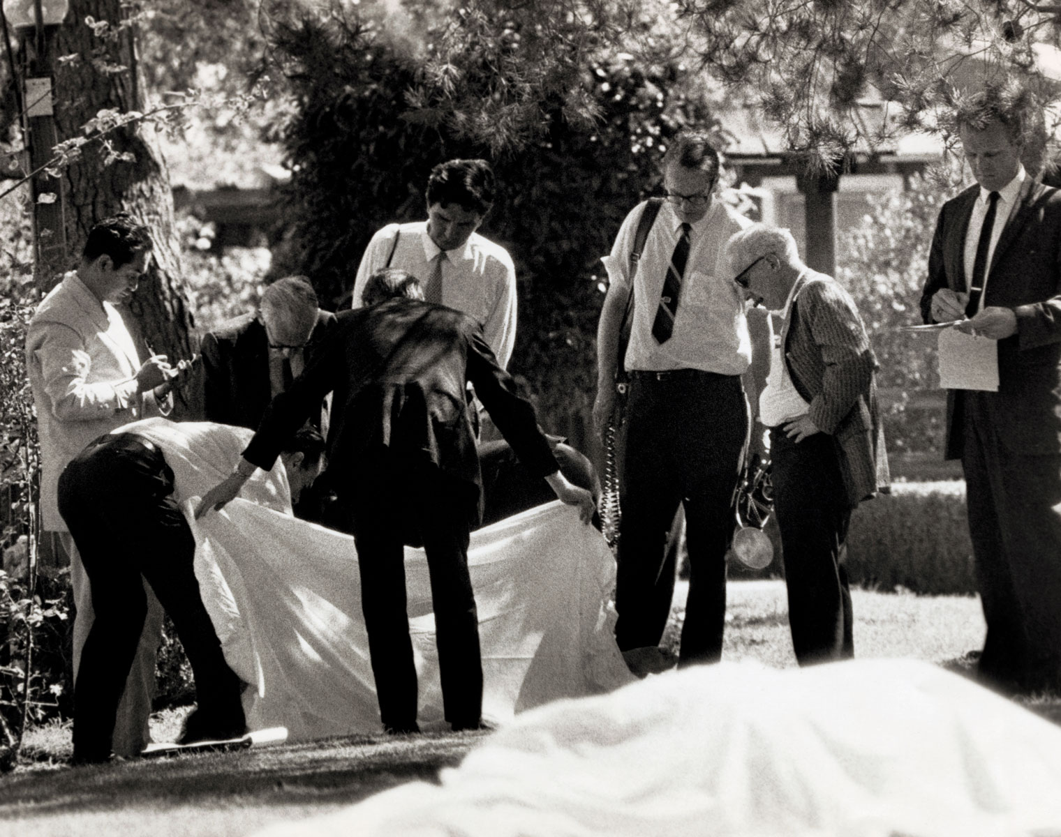 Тело одной из жертв секты Чарльза Мэнсона County Coroner Thomas Nogouchi (left) takes notes as assistants hold sheet which covered one body as the other of the two bodies found on the lawn of the Sharon Tate-Roman Polanski home shows in foreground. Five persons were discovered murdered on the property early August 9th, the work of the Manson Family.